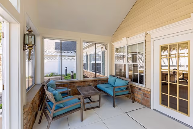 A bright enclosed patio area with large windows and a glass door. The space features two wooden chairs and a wooden loveseat with blue cushions, arranged around a small wooden coffee table. The walls are light-colored with stone accents along the lower half, and the floor is tiled in a light color.
