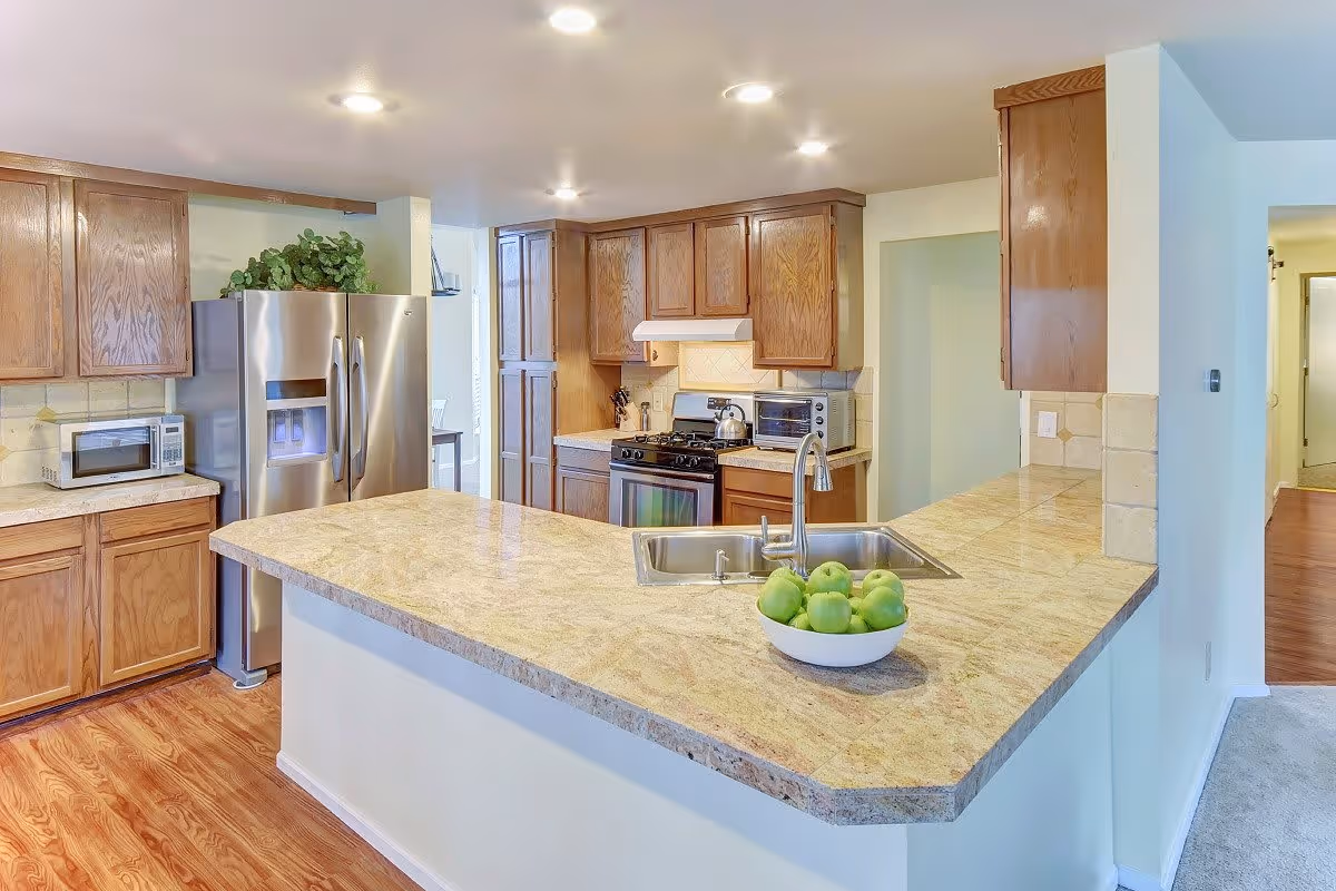 A modern kitchen with wooden cabinets, a stainless steel refrigerator, a microwave, a stove with an oven, and a countertop with a sink. A bowl of green apples is placed on the countertop. The kitchen has recessed lighting and a wooden floor.