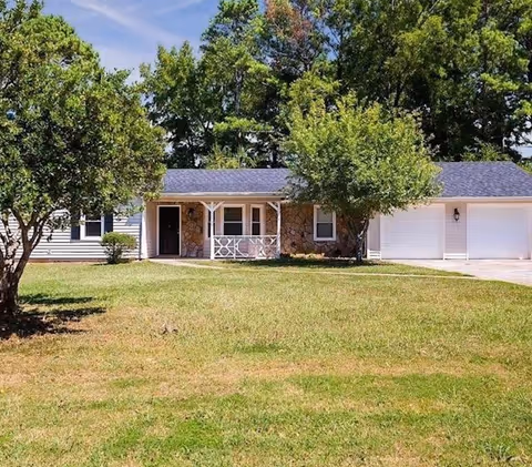 Single-story residential building with a front porch, two garage doors, and a grassy lawn with trees in the foreground under a blue sky.