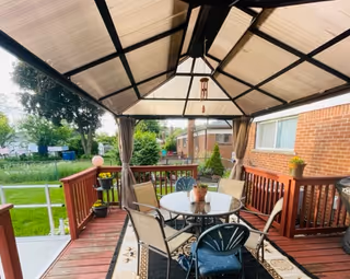Covered wooden deck with a gazebo containing a round glass dining table, chairs, potted plants, and a view of a grassy yard beside a brick building.