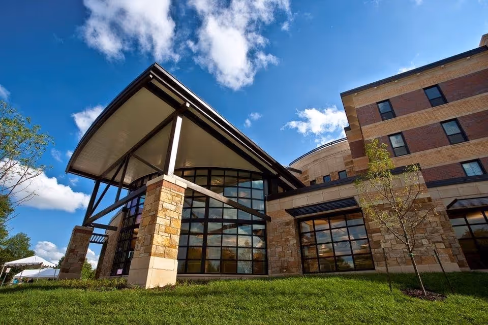 Exterior view of a modern building with large glass windows, stone and brick walls, and a distinctive curved roof structure under a blue sky with scattered clouds. There is green grass and a small tree in the foreground.