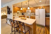 A kitchen area in a senior living facility featuring wooden cabinets, a white refrigerator, microwave, and stove. There is a kitchen island with a light-colored countertop and three decorative chairs. The floor is wood, and the kitchen opens into a hallway with a glimpse of a bedroom.