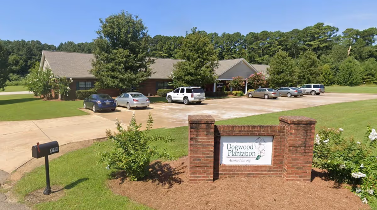 Exterior view of Dogwood Plantation Assisted Living facility with a brick sign in the foreground, a parking lot with several cars, and a single-story building surrounded by trees and greenery under a clear blue sky.