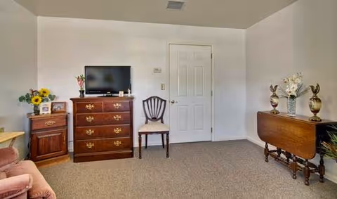 Living room with a TV on a wooden dresser, a chair and side tables against a plain white wall.