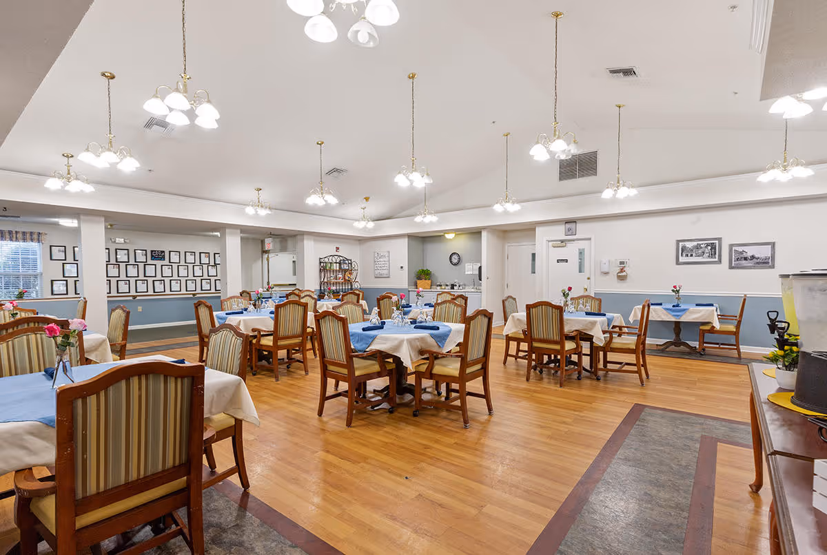 A spacious dining room with multiple tables covered in white and blue tablecloths, each set with glasses and small flower vases. The room has wooden floors, striped cushioned chairs, and several hanging light fixtures. The walls are decorated with framed pictures and a clock, and there is a beverage station on the right side.
