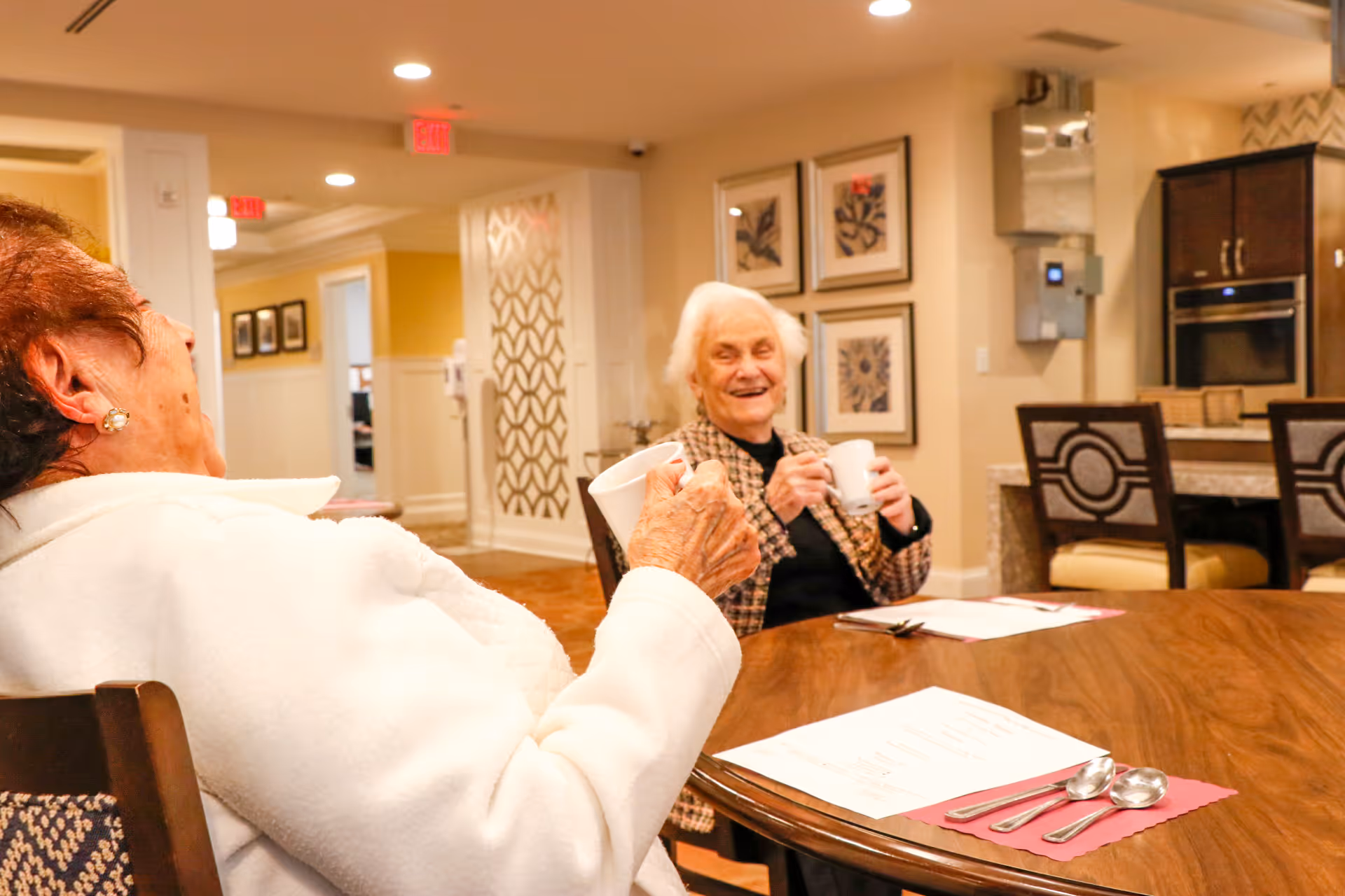 Two elderly women sitting at a wooden table in a well-lit room, smiling and holding white mugs. The room has framed artwork on the wall, a kitchen area with an oven and chairs in the background, and menus with silverware placed on the table.