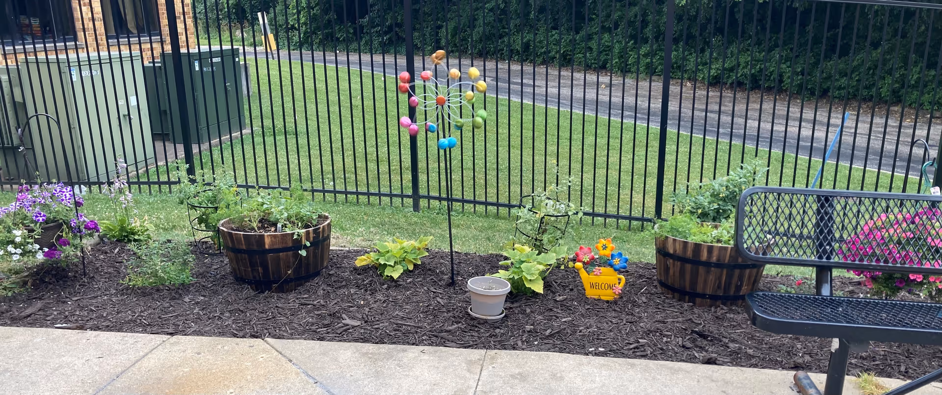 A small garden area with flower pots and plants arranged along a mulched bed next to a black metal fence. There is a colorful pinwheel decoration in the center and a yellow watering can with a welcome sign. A black metal bench is partially visible on the right side, and a grassy area and paved path are seen beyond the fence.