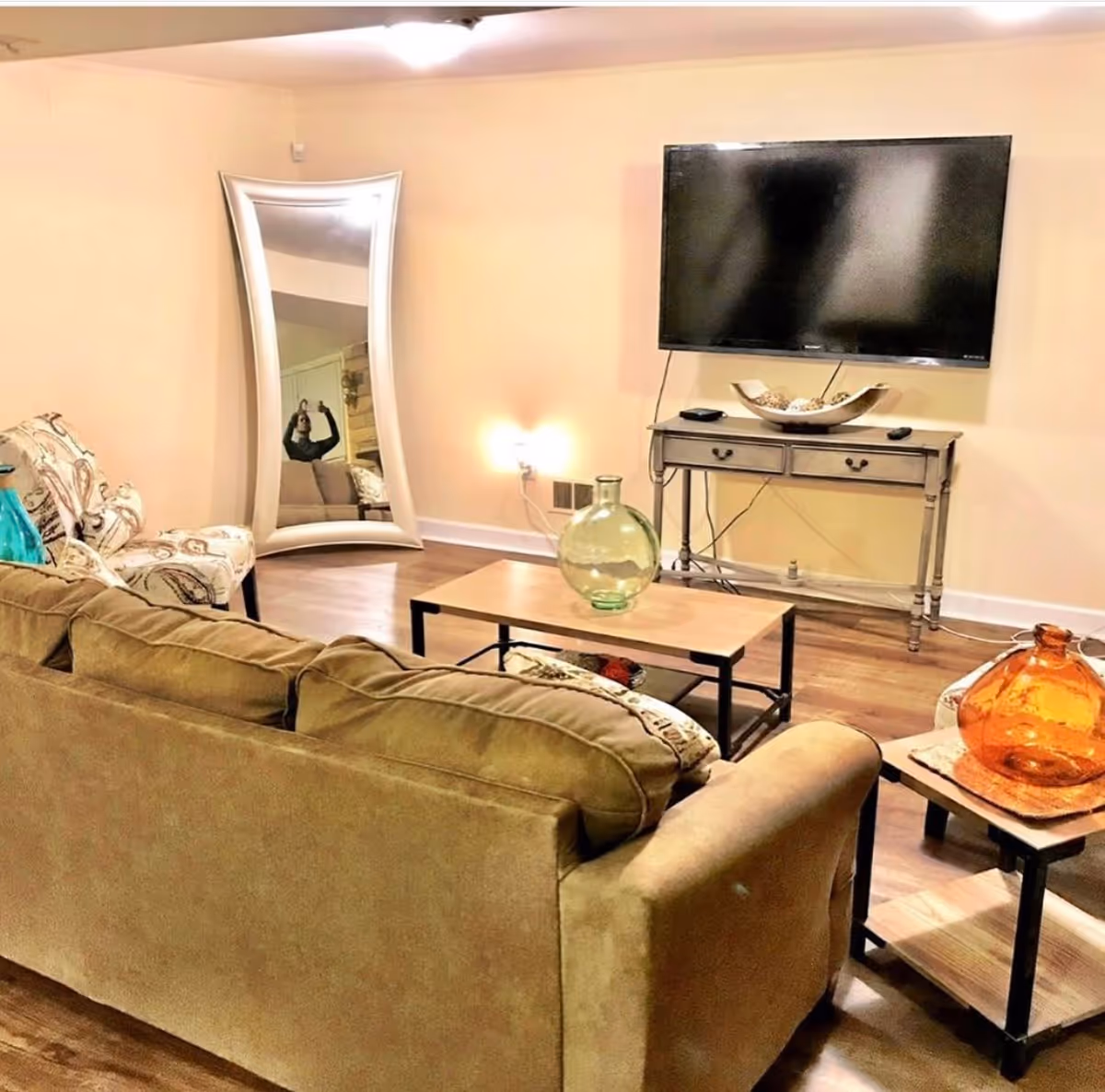 A cozy living room with a beige sofa, patterned armchair, wooden coffee table with a green glass vase, side table with an orange glass vase, a large wall-mounted flat screen TV above a small console table, and a tall curved mirror leaning against a beige wall.