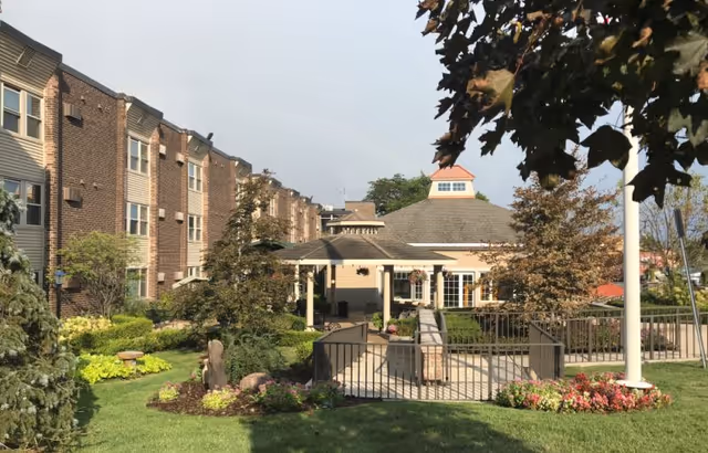 Outdoor view of The Bellaire Senior Living facility showing a landscaped garden with green grass, trees, and flower beds. There is a paved walkway leading to a covered seating area attached to a multi-story brick and siding building under a clear sky.