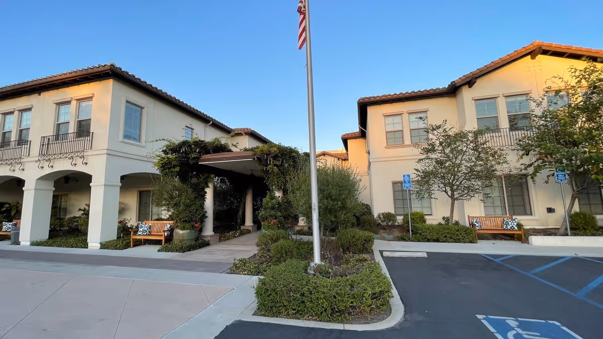 Front entrance of a Mediterranean-style senior living building with a flagpole, benches, landscaping, and accessible parking spaces.