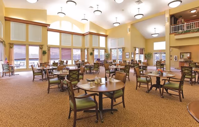 A spacious dining room with multiple round tables and chairs arranged neatly. The room has large windows with blinds, letting in natural light. The walls are painted a warm yellow, and there are plants and framed pictures decorating the space. The ceiling is high with several round light fixtures, and there is a balcony area visible on the right side.