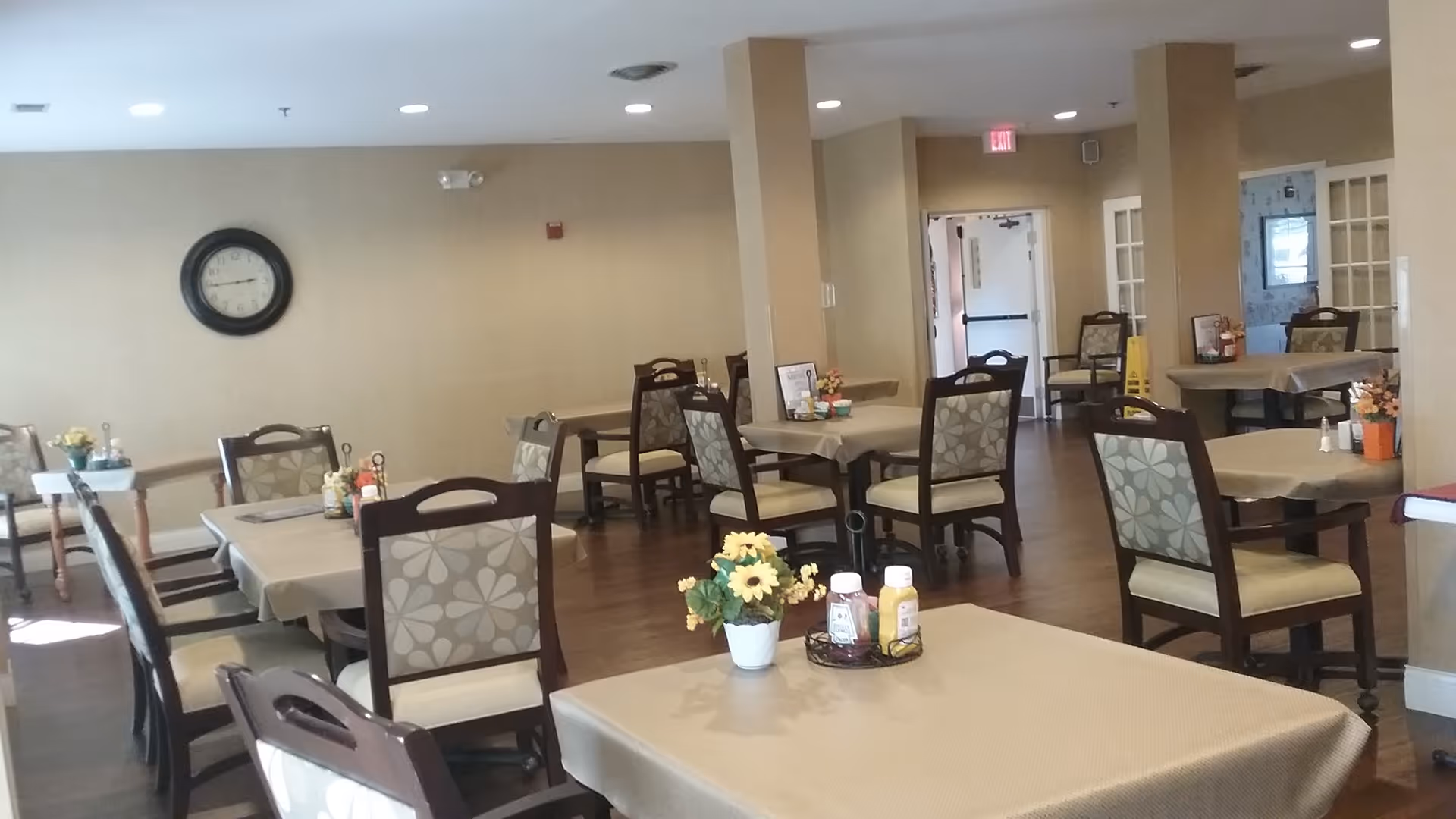 A dining room with multiple tables covered with beige tablecloths, each surrounded by chairs with floral patterned upholstery. Small flower arrangements and condiments are placed on the tables. The room has beige walls, wooden flooring, and a clock on one wall. There is an exit door visible in the background.