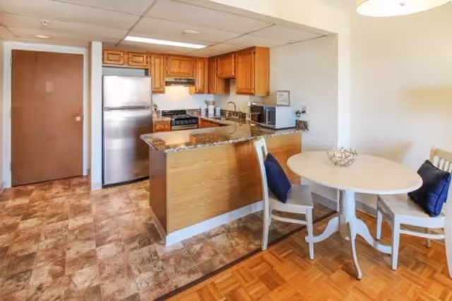A kitchen and dining area in a senior living community. The kitchen features wooden cabinets, a stainless steel refrigerator, a stove with an overhead vent, a microwave, and a granite countertop with a breakfast bar. Adjacent to the kitchen is a small round white dining table with two chairs, each with a dark blue cushion. The floor is a combination of tiled and parquet wood.