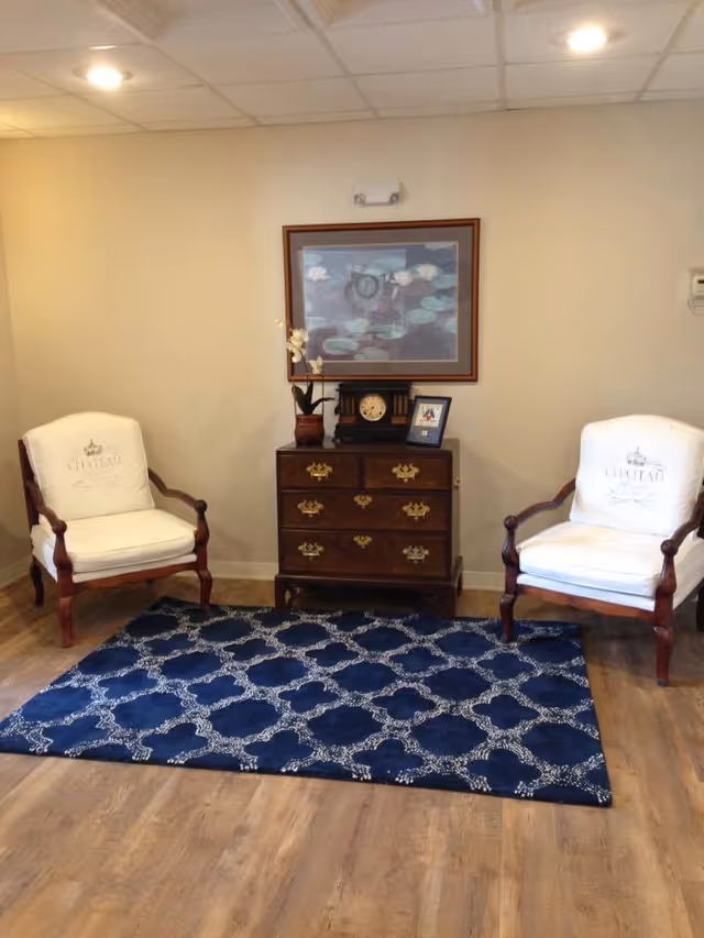 A cozy sitting area with two white cushioned armchairs with wooden frames placed on either side of a small wooden chest of drawers. On top of the chest is a decorative clock, a potted orchid, and a framed photo. Above the chest hangs a framed painting of water lilies. The floor has a blue patterned rug, and the walls are painted light beige.