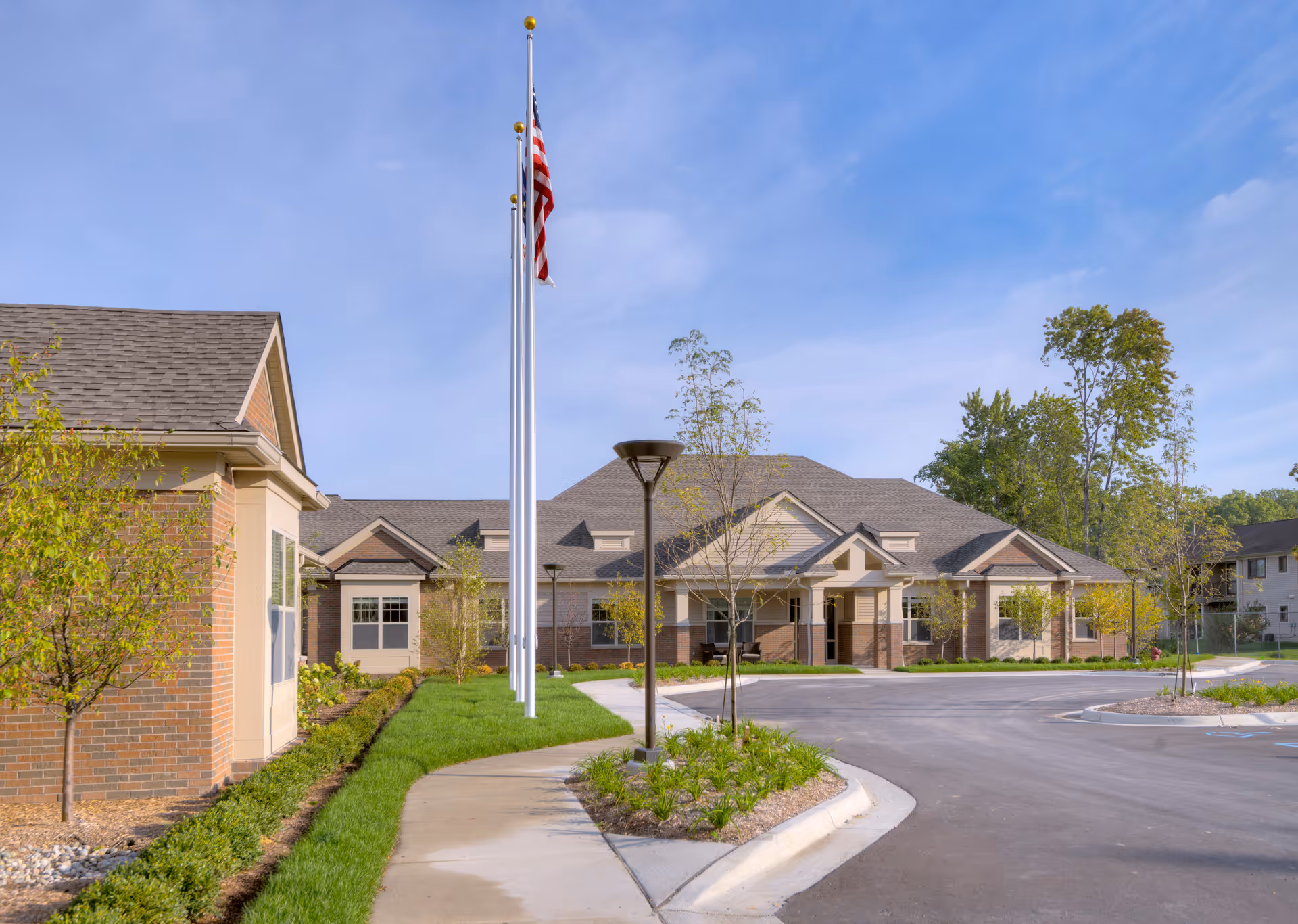 Exterior view of a single-story senior living facility building with a paved driveway, landscaped greenery, young trees, and three flagpoles with an American flag flying. The sky is clear and blue.