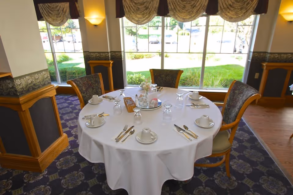 Round dining table set with white tablecloth, cups, saucers, utensils, and upside-down glasses in a room with large windows showing a green outdoor area. Four upholstered chairs surround the table, and the room has patterned carpet and decorative wall paneling.