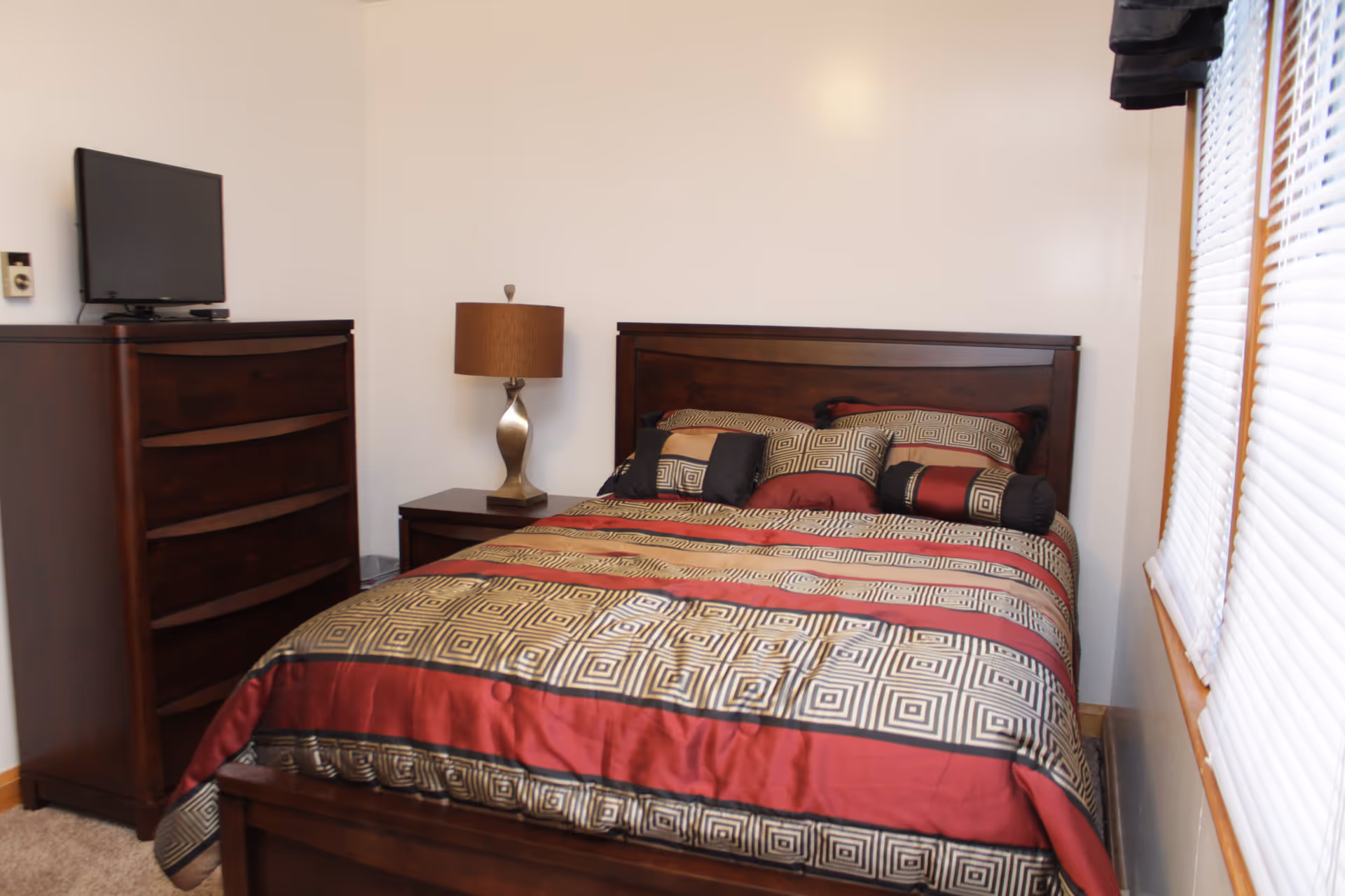 A bedroom with a wooden bed frame and a bed made with a patterned comforter and multiple pillows. Next to the bed is a wooden nightstand with a modern lamp. Across from the bed is a tall wooden dresser with a flat-screen TV on top. A window with white blinds is on the right side of the room.
