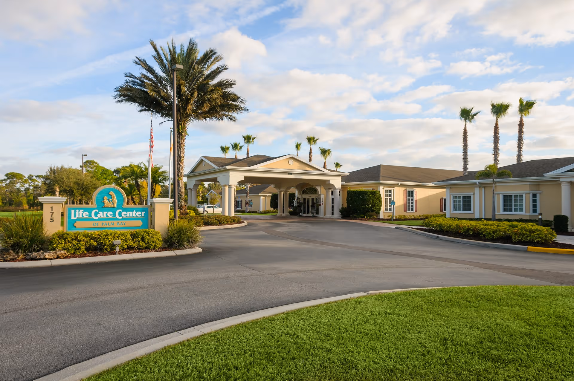 Front entrance of the Life Care Center of Palm Bay with a driveway, landscaped islands, entrance canopy, and palm trees.