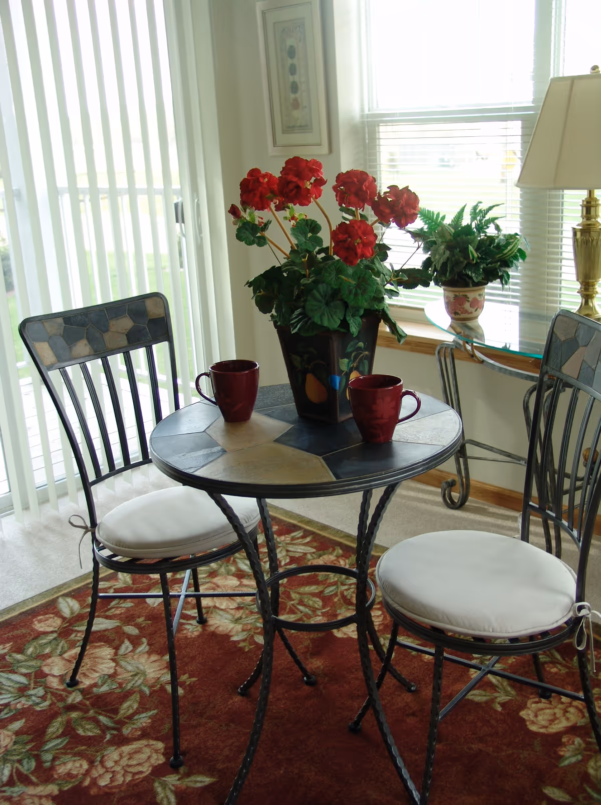 Small round bistro table with two chairs, a potted red-flowered centerpiece and two mugs in a sunny windowed dining nook.