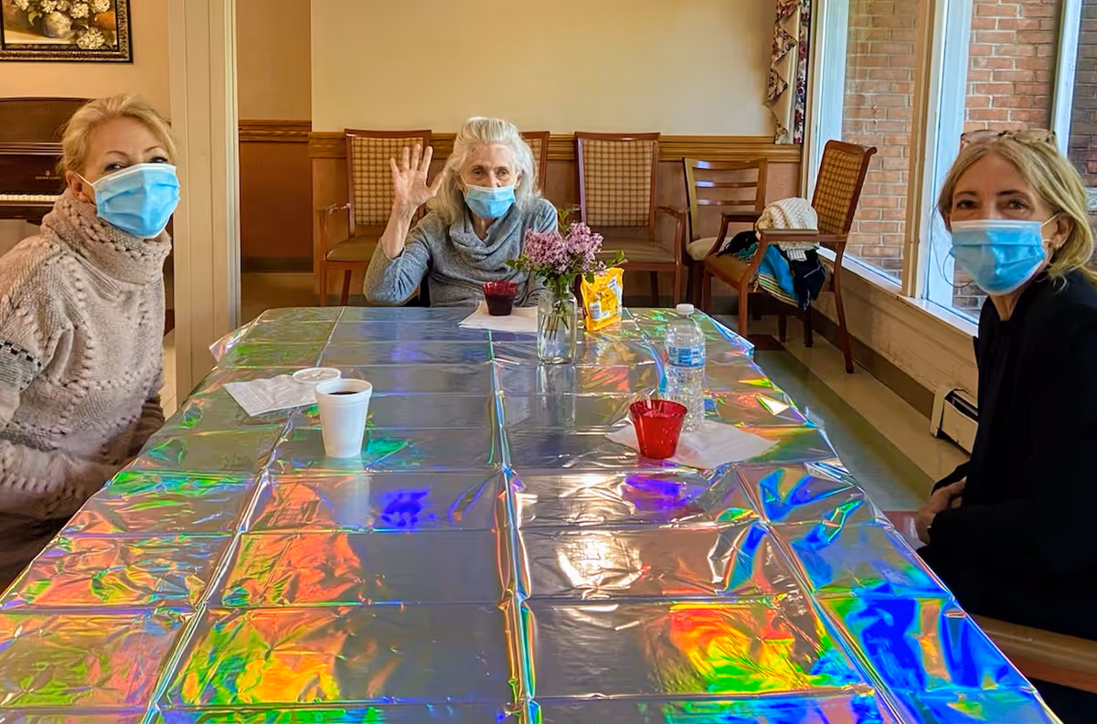 Three women wearing face masks sitting around a table covered with a shiny, reflective tablecloth. One woman in the center is waving. There are chairs and a window with a brick wall outside in the background. A vase with flowers and some cups and bottles are on the table.