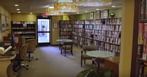 Interior library/reading room with rows of bookshelves, round tables and chairs, and a doorway at the back.