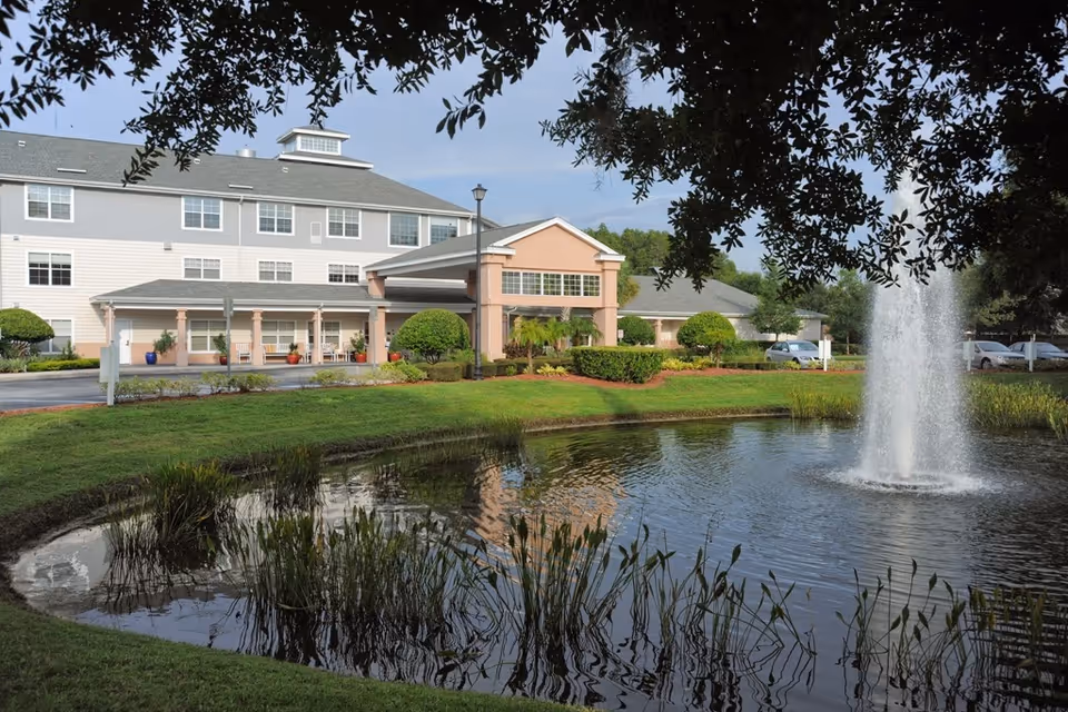 Exterior view of a multi-story senior living facility building with a covered entrance, surrounded by well-maintained landscaping including trimmed bushes and a pond with a water fountain in the foreground. Trees frame the top of the image.