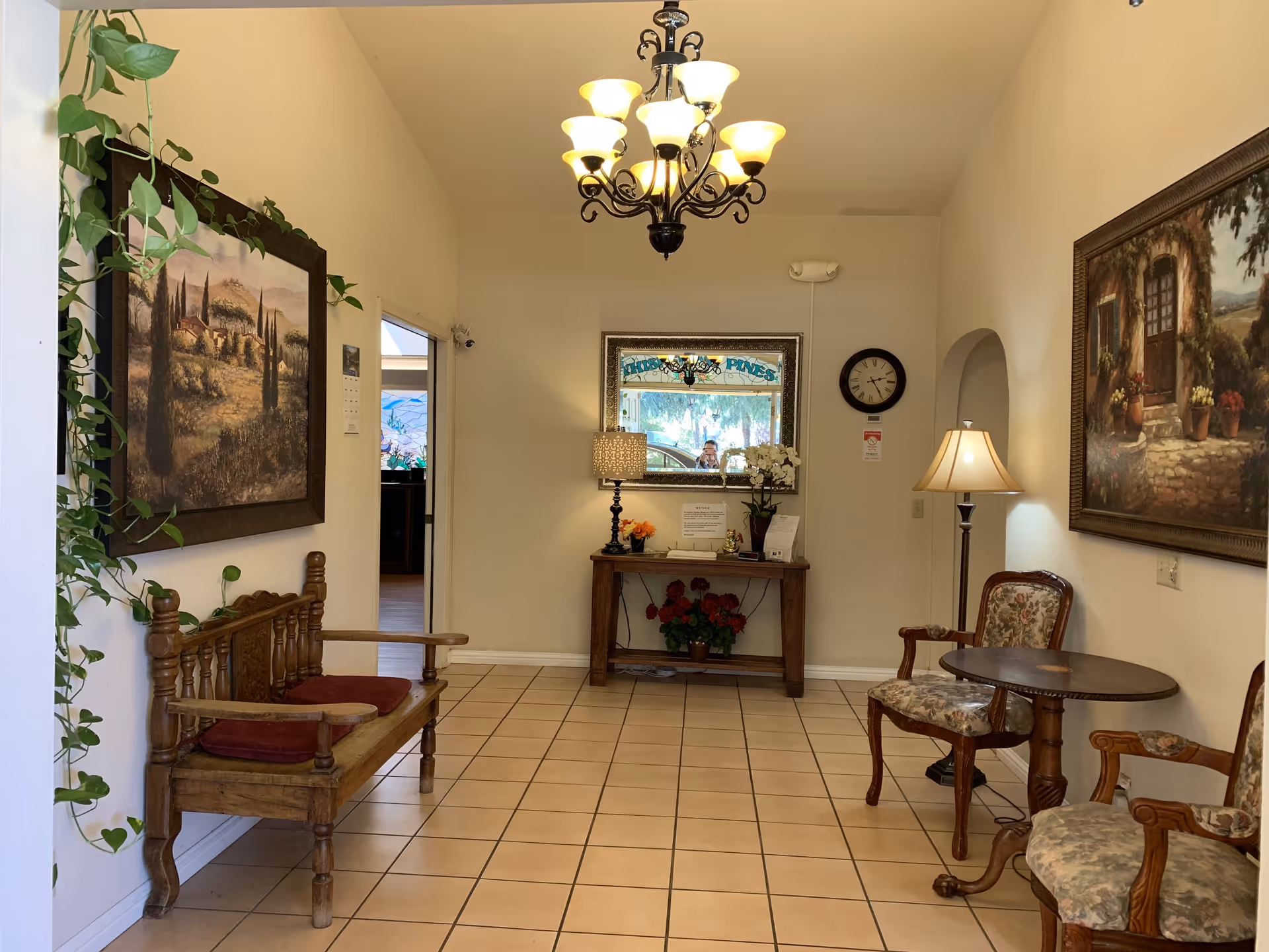 A cozy interior seating area with a wooden bench with red cushions on the left, two floral upholstered armchairs with a round wooden table on the right, and a wooden console table with a lamp, flowers, and a mirror on the back wall. The room has beige tiled flooring, cream-colored walls, a chandelier hanging from the ceiling, and framed paintings on the walls.