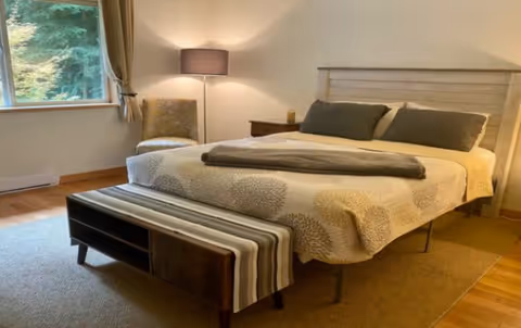 A cozy bedroom with a large bed covered in a beige and gray patterned quilt, two gray pillows, and a folded blanket at the foot. There is a wooden bench with a striped cushion at the foot of the bed. A cushioned chair and a floor lamp are positioned near a window with beige curtains, allowing natural light to enter the room.
