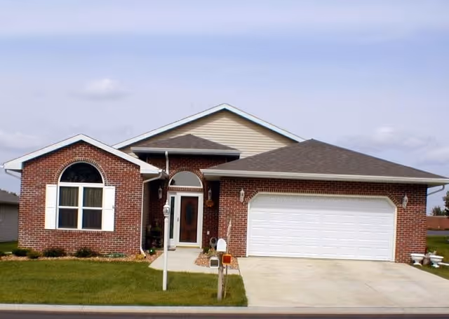 Front exterior of a single-story brick house with an attached two-car garage, driveway, and small lawn.