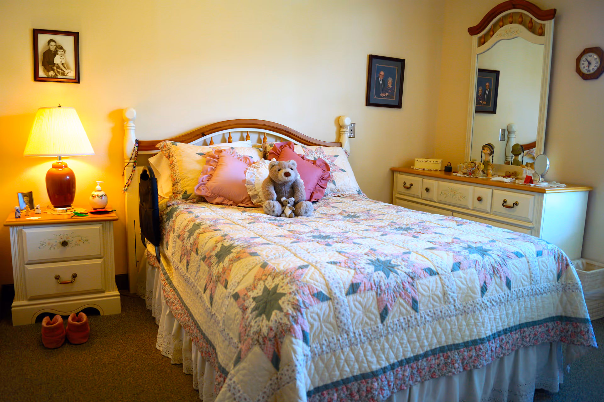A cozy bedroom with a bed covered in a colorful quilt featuring star patterns. The bed has multiple pillows and a teddy bear placed on top. To the left of the bed is a nightstand with a red lamp, small framed photo, and other small items. On the right side, there is a dresser with a large mirror and various decorative items. The walls are light-colored with framed pictures hanging above the bed and dresser. A pair of slippers is placed on the floor near the nightstand.