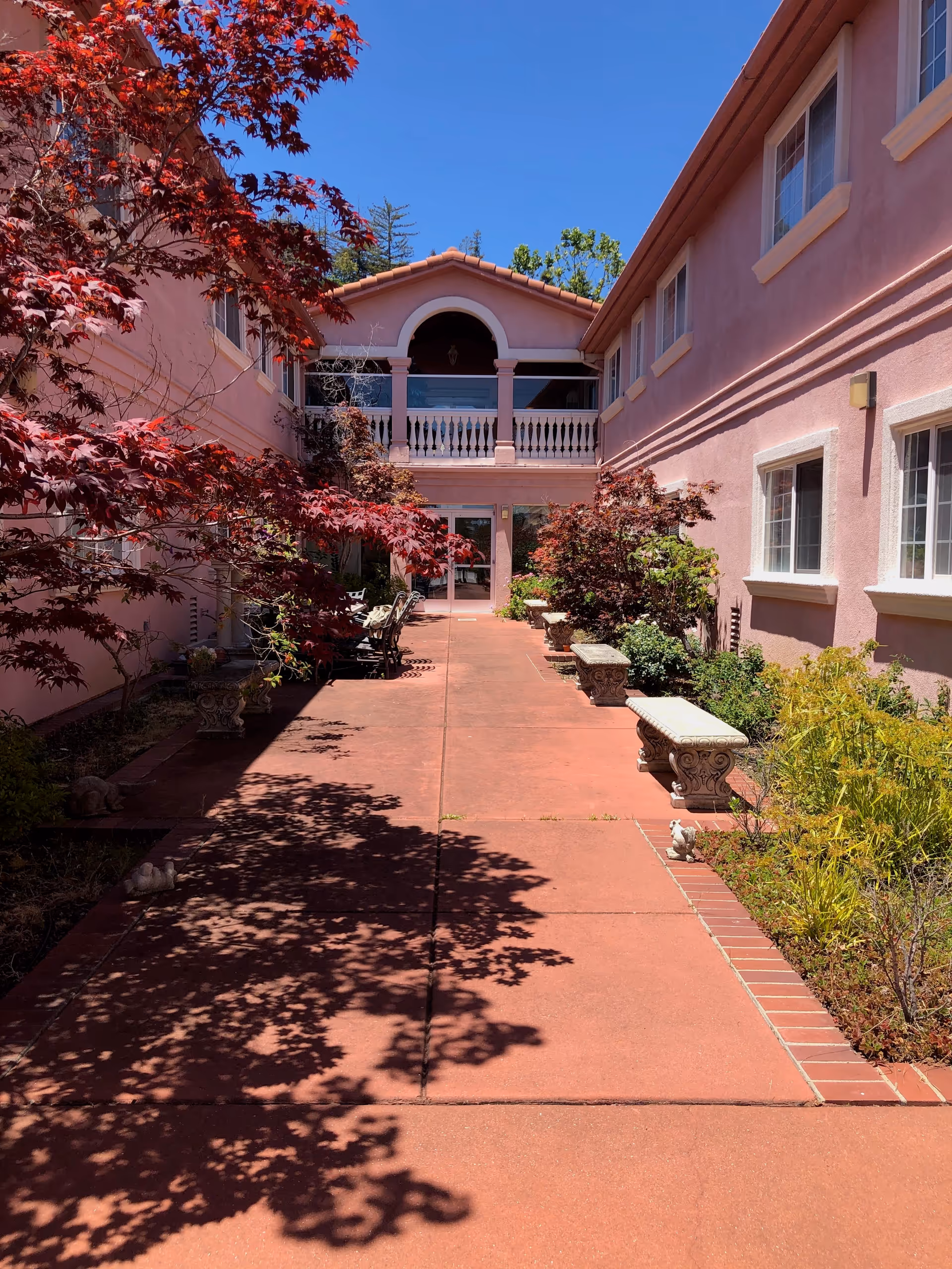 Outdoor courtyard area of a senior living facility with pink stucco walls, red tiled roof, and a paved walkway. The courtyard is lined with benches, plants, and small trees with red and green foliage under a clear blue sky.