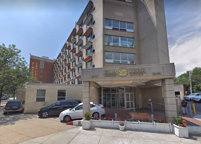 Exterior view of Elm York Assisted Living facility showing a multi-story building with a covered entrance, several parked cars, and some greenery around the entrance area.