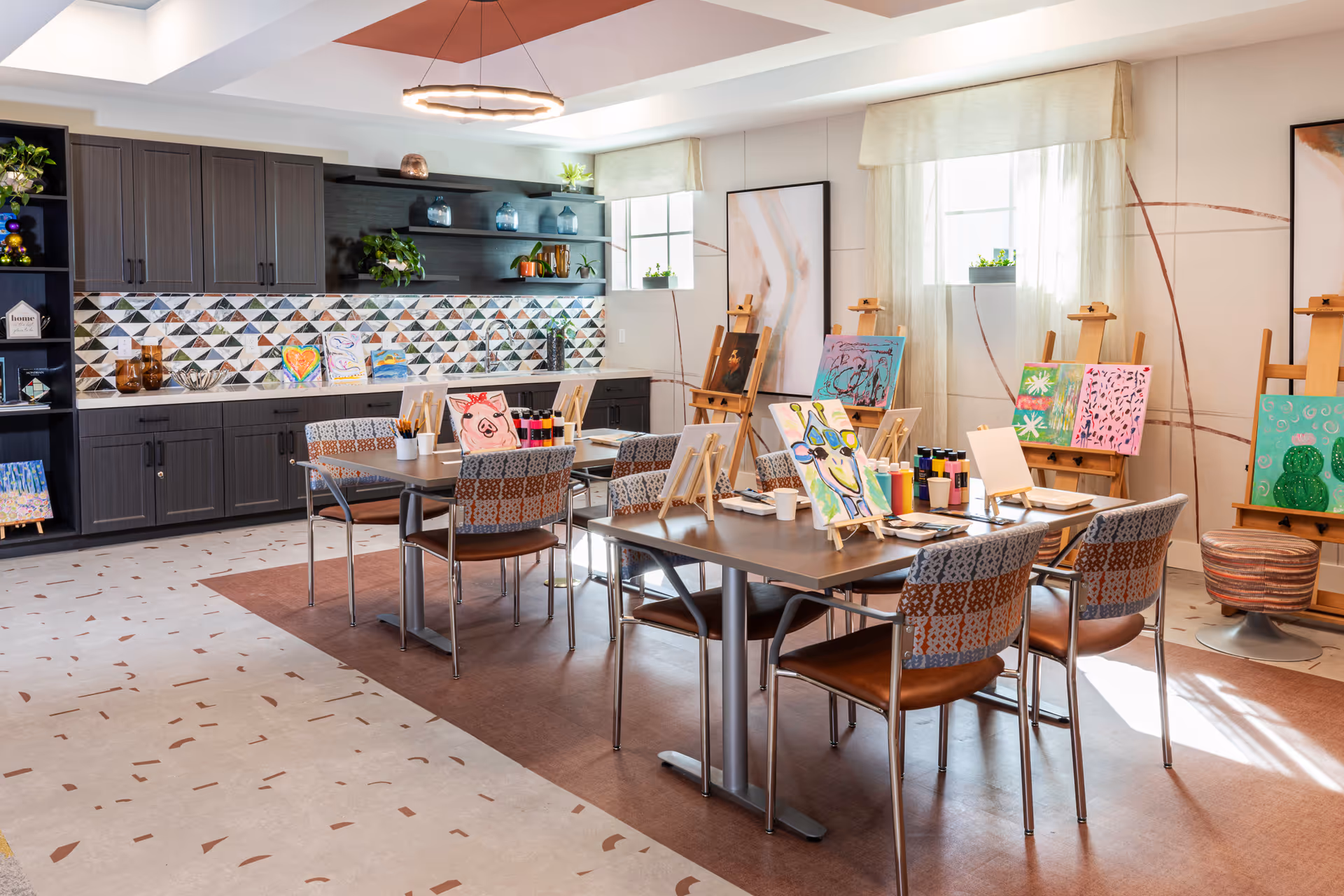 A bright and inviting art room with two tables and several chairs arranged around them. Each table has easels holding colorful paintings, paint bottles, brushes, and paper cups. The room features a modern design with patterned floor tiles, a geometric tiled backsplash, dark cabinetry, open shelves with decorative items, and two windows with sheer curtains letting in natural light.