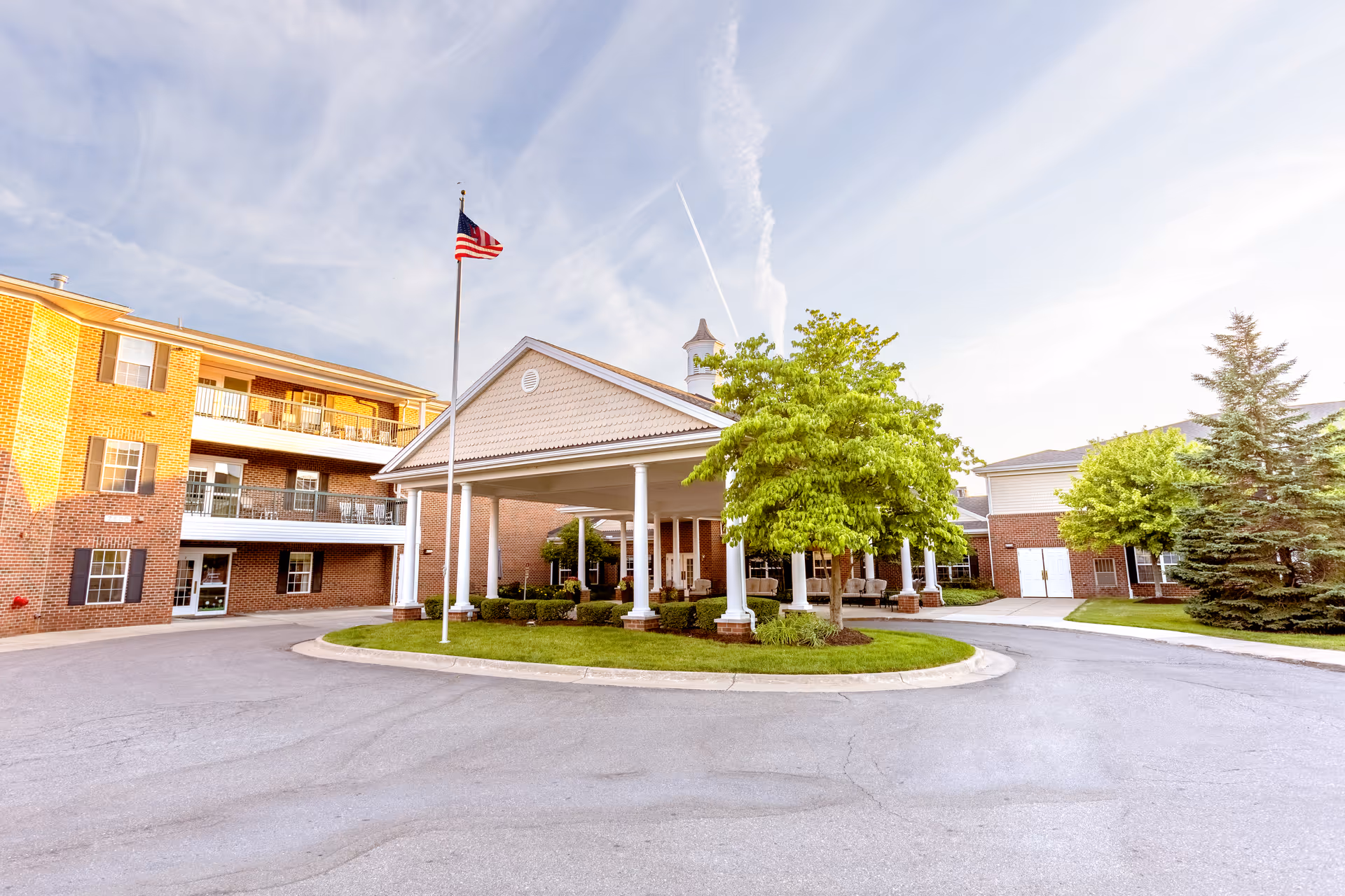 Exterior view of Independence Village of Brighton Valley showing a covered entrance with white columns, a circular driveway, an American flag on a flagpole, and surrounding greenery including trees and shrubs. The building is made of brick with multiple windows and balconies.