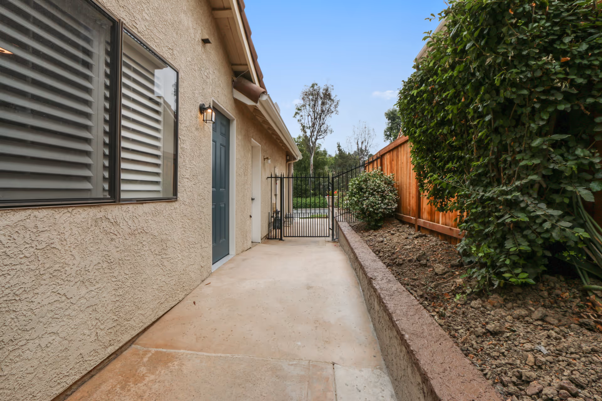 Outdoor walkway beside a beige stucco building with a blue door and a window with blinds. The walkway is paved and bordered by a raised garden bed with soil and green bushes. A black metal gate is visible at the end of the walkway, with a wooden fence and trees in the background under a clear blue sky.