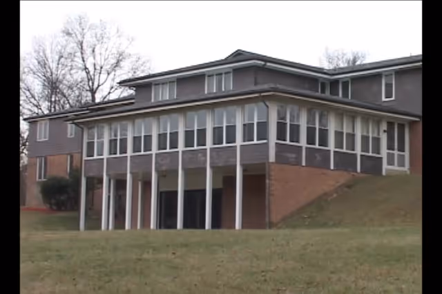 Exterior view of a multi-story assisted living facility building with large windows and white pillars supporting an extended upper floor. The building is surrounded by grass and leafless trees under an overcast sky.