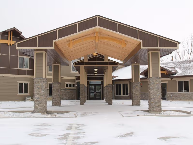 Front exterior view of Mountain Lodge LLC building with a covered entrance supported by stone pillars, surrounded by snow-covered ground and a cloudy sky.