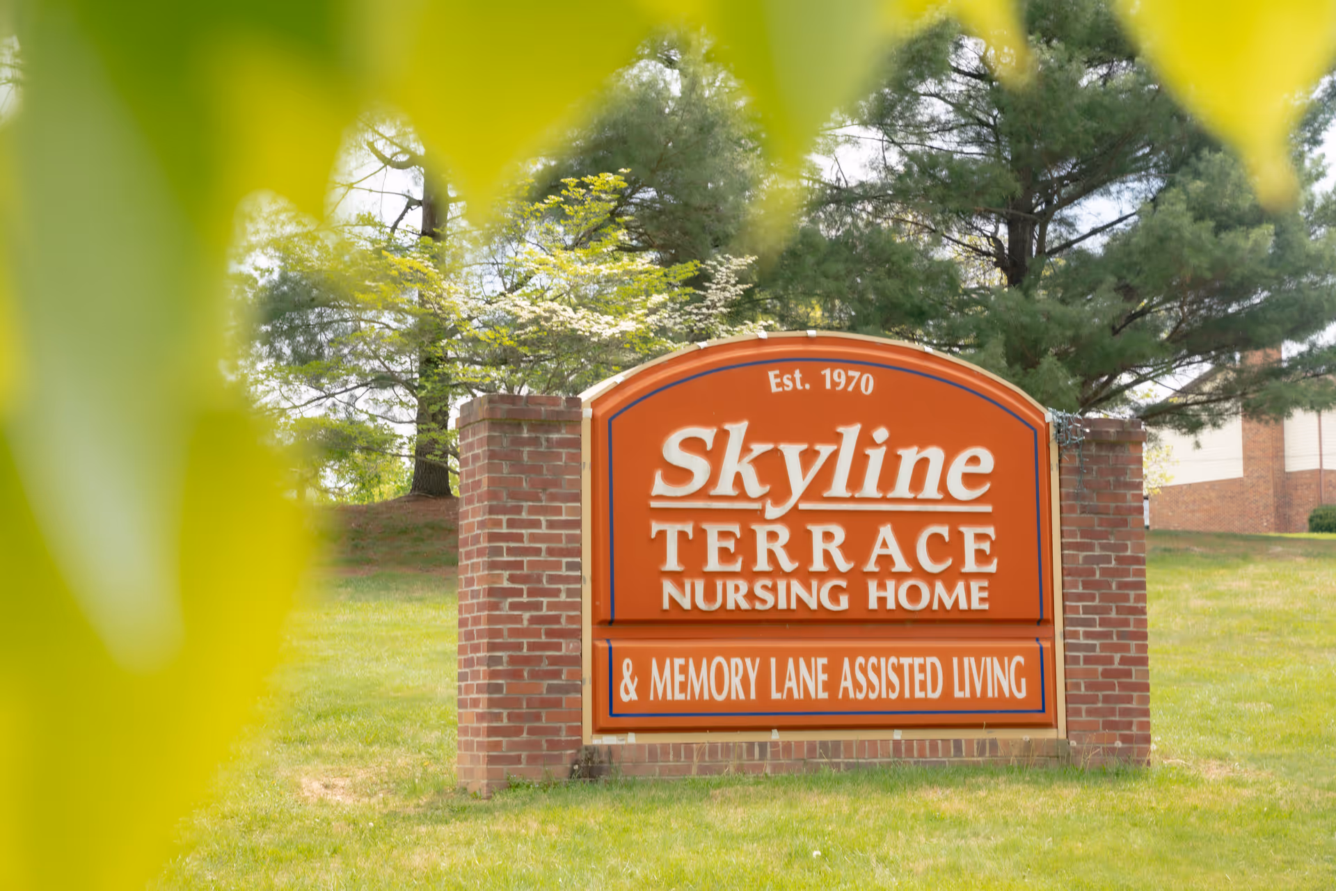 Outdoor brick sign for Skyline Terrace Nursing Home & Memory Lane Assisted Living on a grassy lawn with trees and blurred leaves in the foreground.