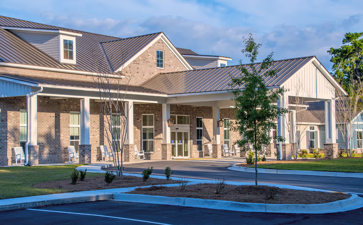 Exterior view of a senior living facility with a covered entrance, brick walls, metal roof, several windows, and white rocking chairs along the porch. There is a small landscaped area with a young tree and shrubs in front of the building.