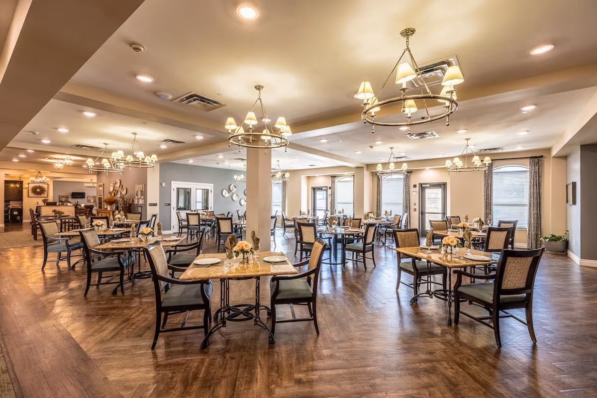 A spacious, well-lit dining room with multiple tables set for meals. Each table has chairs, plates, napkins, and small floral centerpieces. The room features wooden flooring, large windows with curtains, and elegant chandeliers hanging from the ceiling.