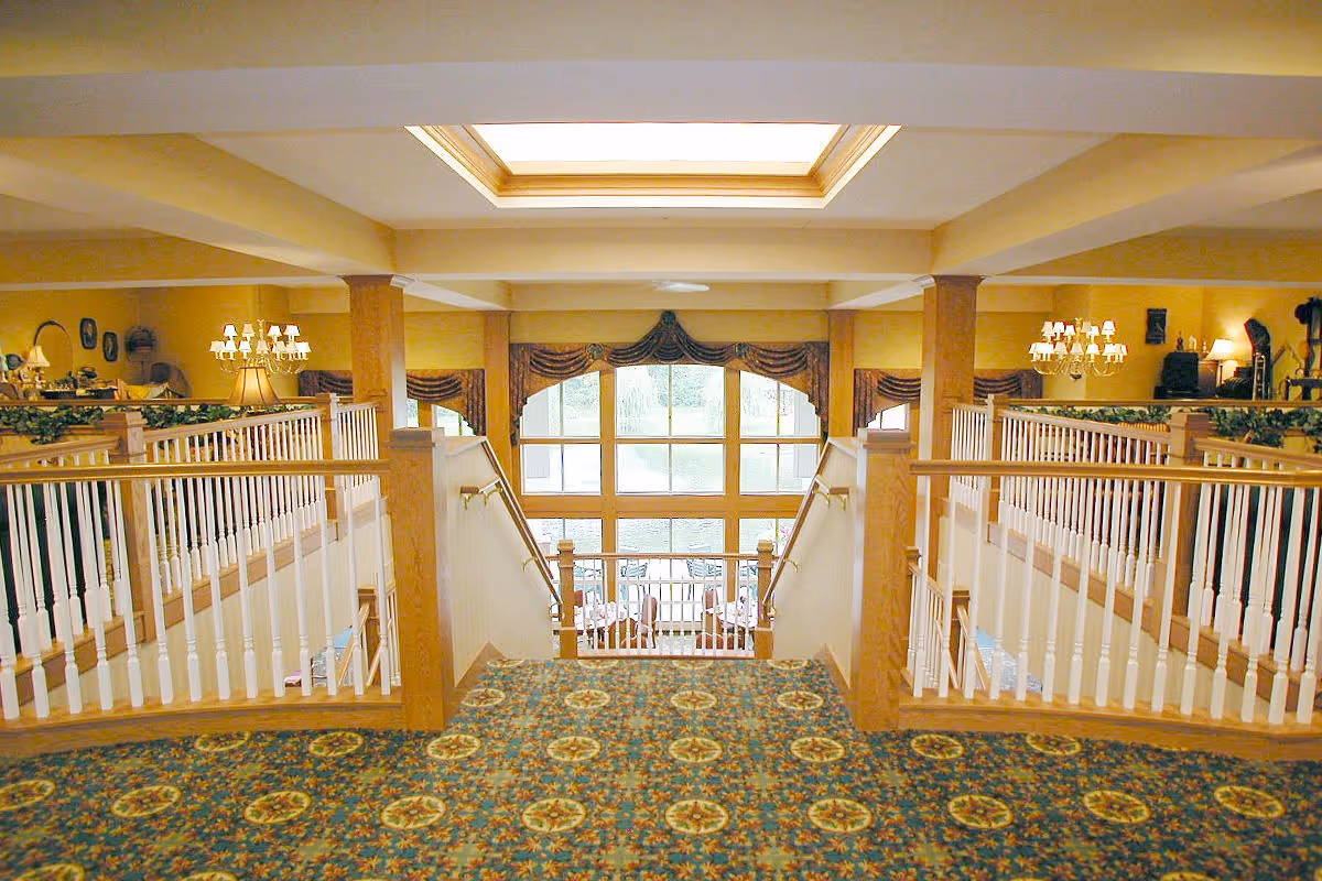Interior view of a senior living facility with a split staircase leading down to a dining area. The space features patterned carpet, wooden railings with white balusters, large windows with draped curtains, and warm yellow walls. Chandeliers and lamps provide lighting, and there is a skylight above.