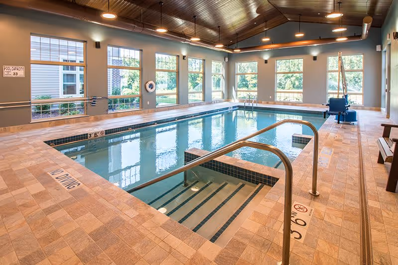 Indoor swimming pool with tiled floor and steps leading into the water. Large windows along the walls let in natural light, and there is a pool lift chair for accessibility. The ceiling has wooden paneling with recessed lighting.