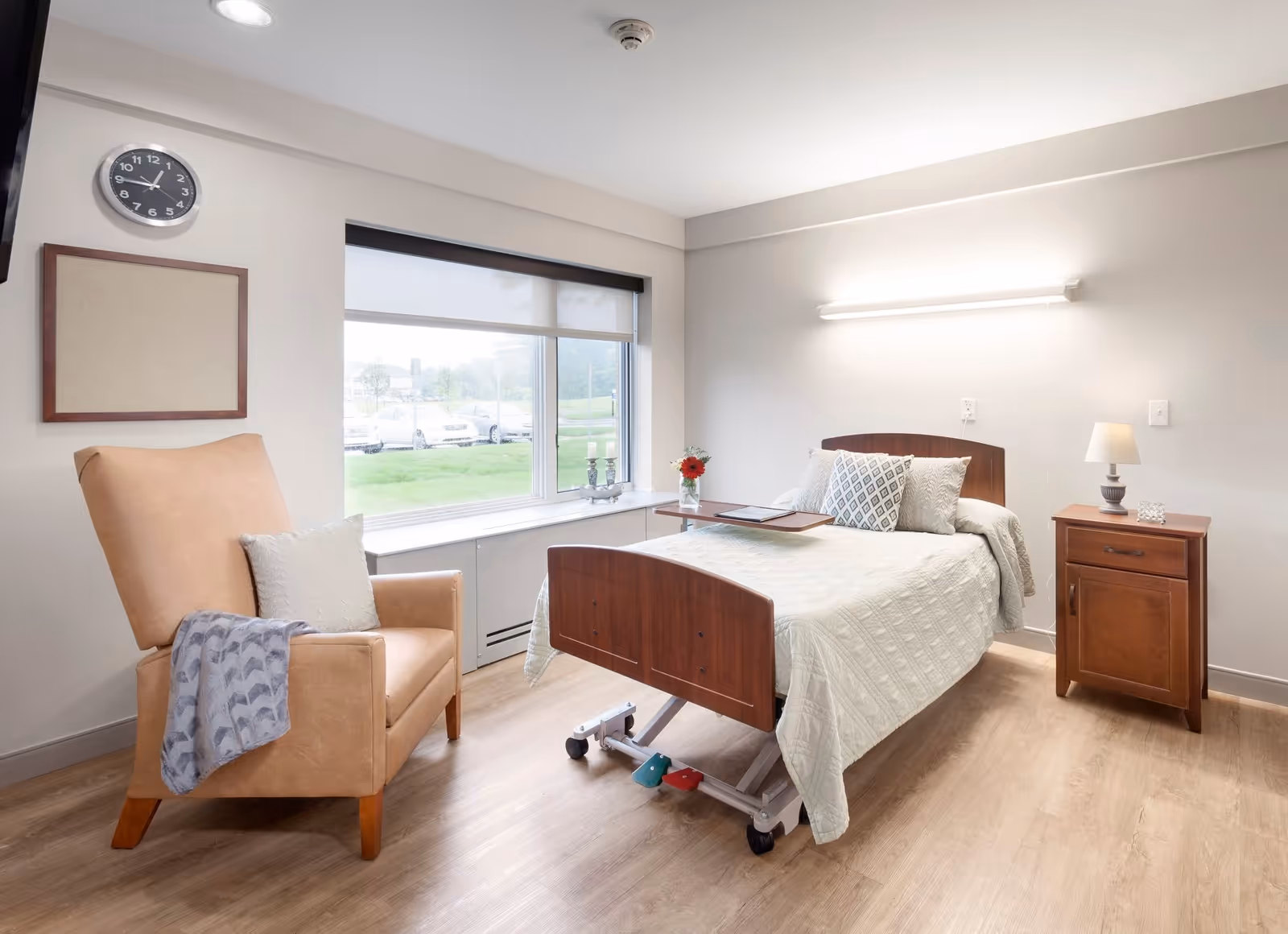 A bright and clean senior living bedroom with a hospital-style adjustable bed covered in a white quilt, a wooden nightstand with a lamp, a beige armchair with a pillow and a blanket, a large window with a view of parked cars and greenery outside, a wall clock, and a blank bulletin board.