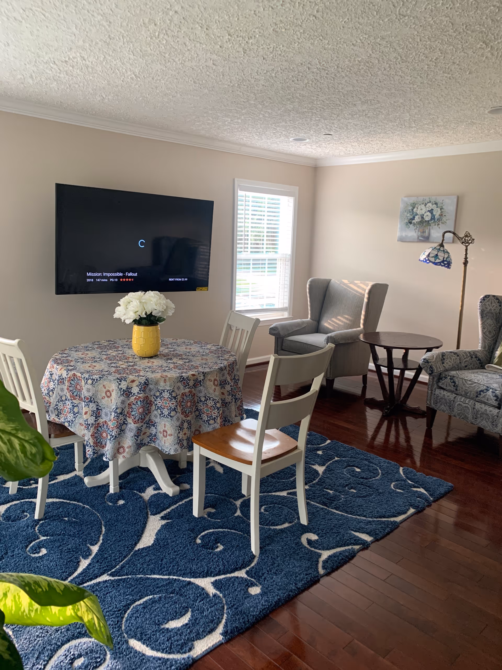Bright living room with a round table covered by a patterned tablecloth and vase of flowers, wall-mounted TV, two armchairs, and a blue patterned rug.