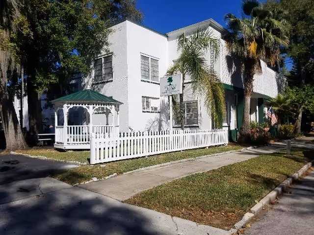 White two-story assisted living facility with a green awning, gazebo, and white picket fence.