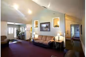A spacious common living area with sofas, lamps, framed artwork, purple carpet and a hallway leading to other rooms.
