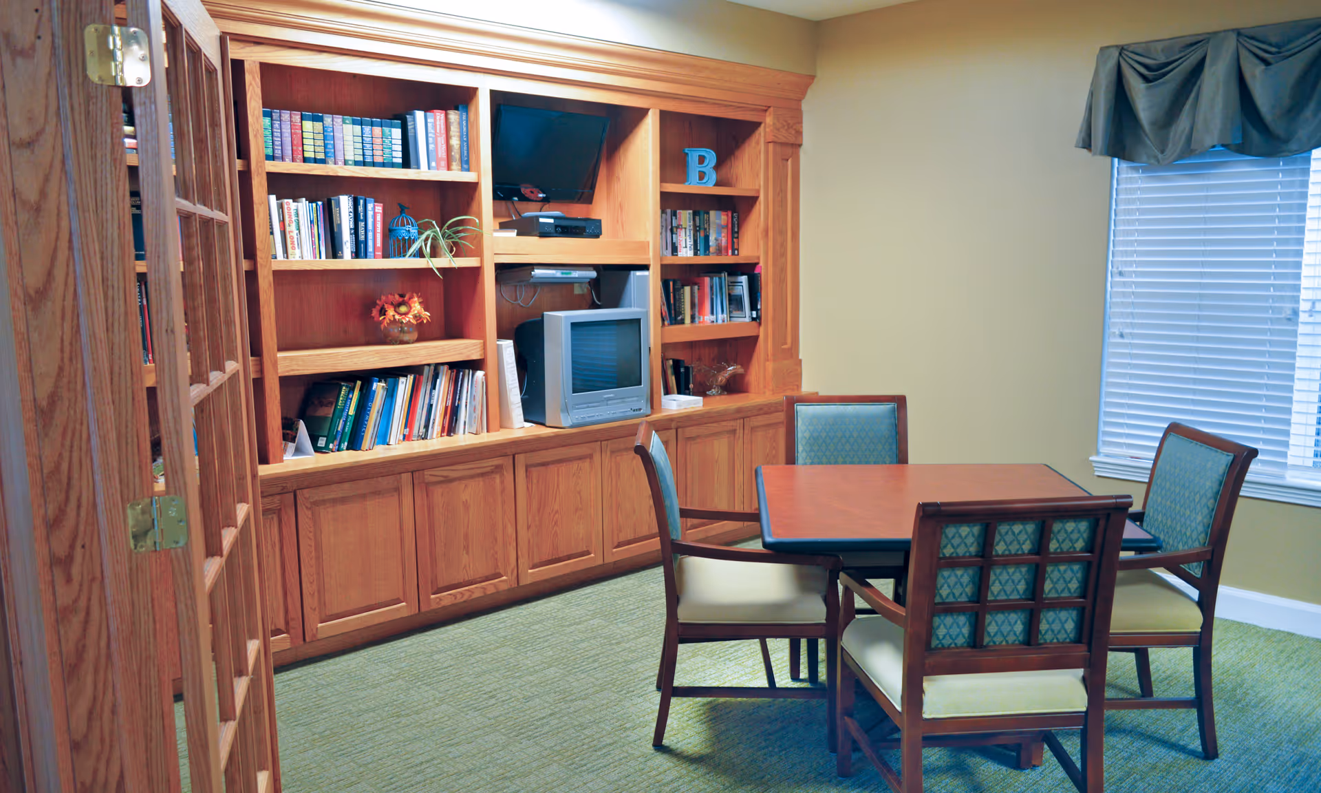 Small common room with built-in wooden bookshelves and a TV, and a square table with four chairs near a window.