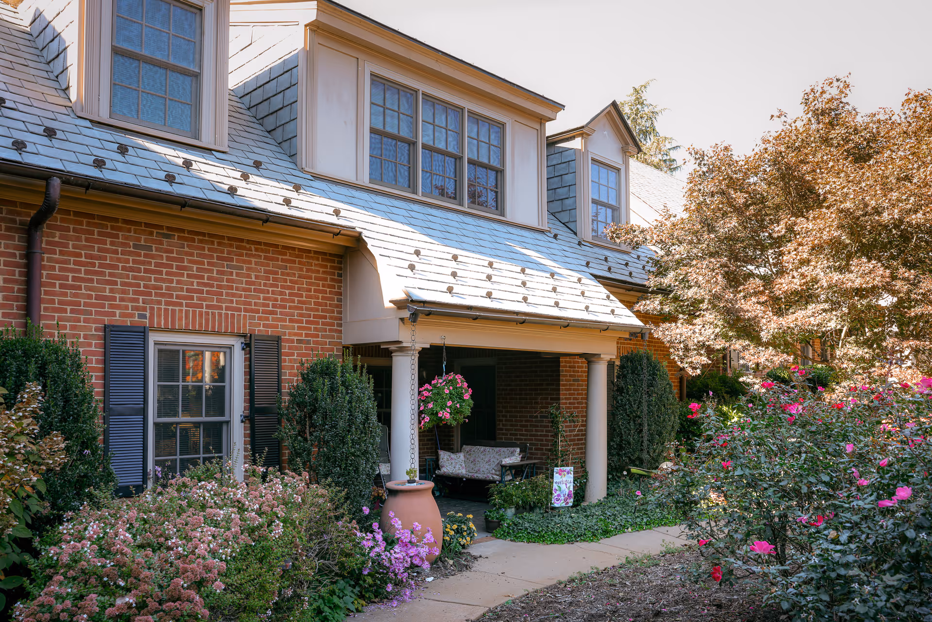 Exterior view of a brick building with a slate roof and dormer windows. The entrance is shaded by a small porch with white columns and a hanging flower basket. The surrounding garden is lush with various flowering plants and shrubs, including pink and purple flowers, and a tree with autumn-colored leaves.