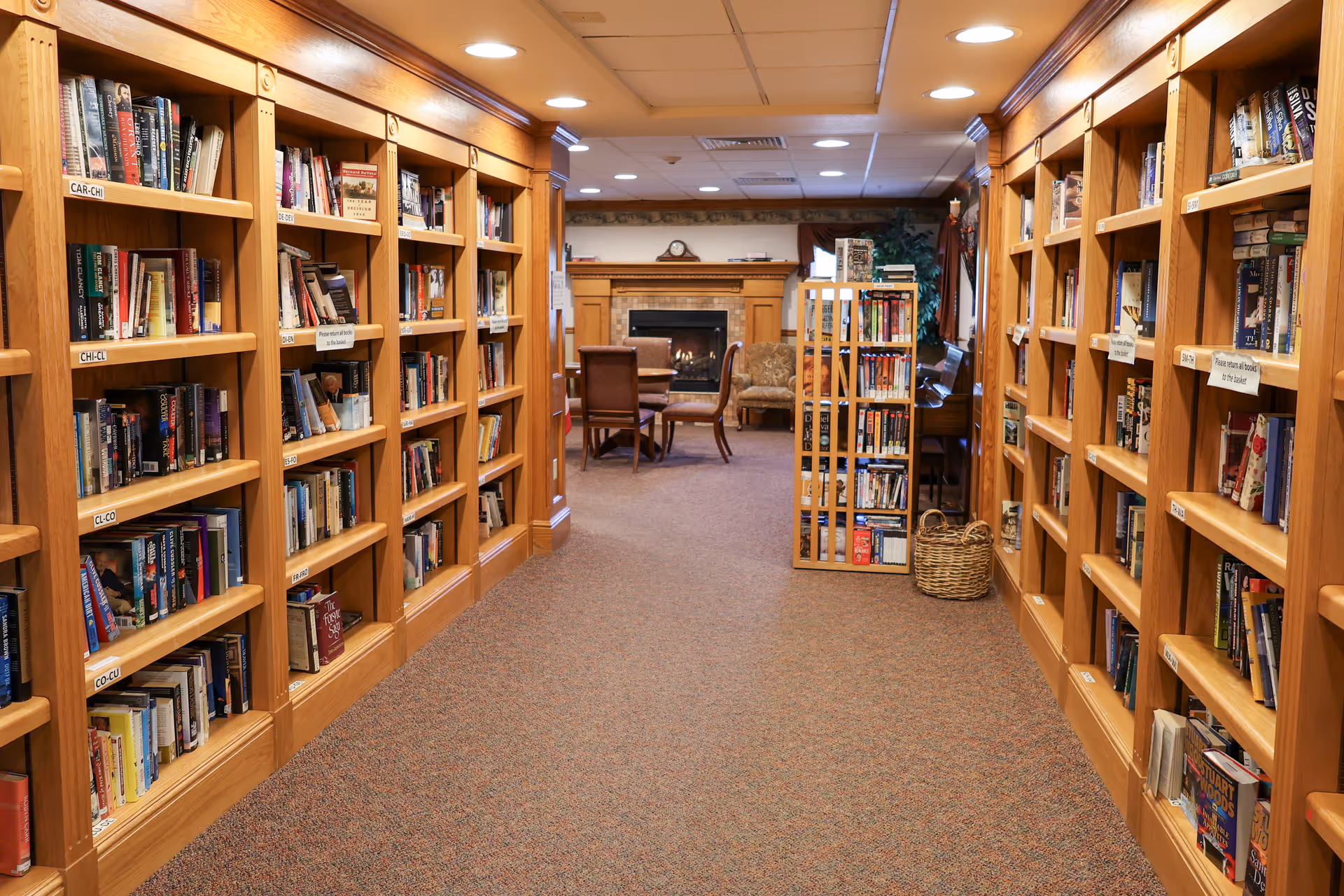 A cozy library room with wooden bookshelves filled with books lining both sides of a carpeted hallway. At the end of the hallway, there is a seating area with chairs and a fireplace, creating a warm and inviting atmosphere.