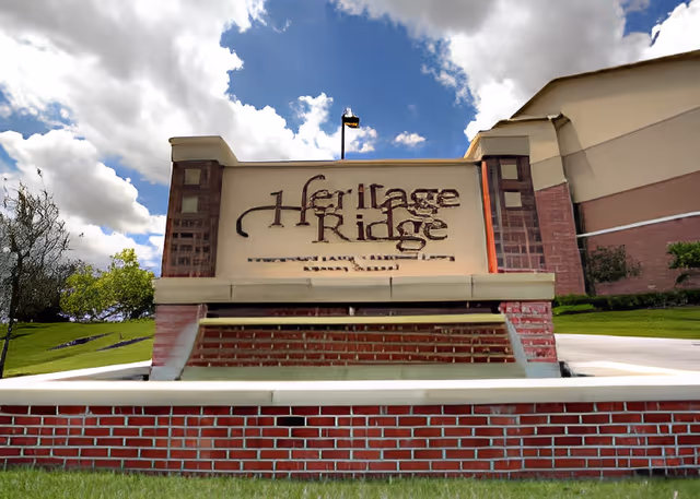 A large brick and stone sign for Heritage Ridge set on a grassy area with a partly cloudy blue sky in the background. The sign is positioned near a building and a flagpole is visible behind it.